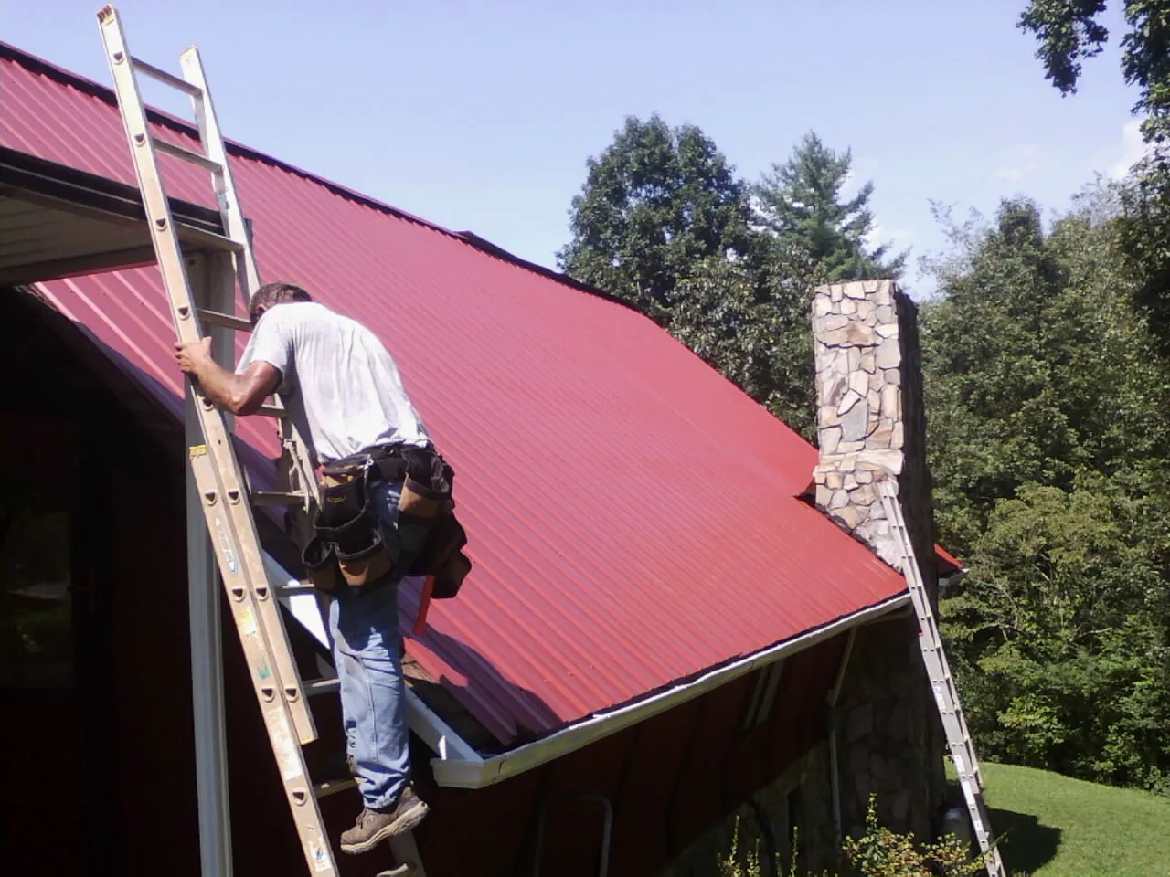 Roofer on a ladder beside a red metal roof and stone chimney — Soffit & Fascia Repair by Roofing Brothers in Bunker Hill